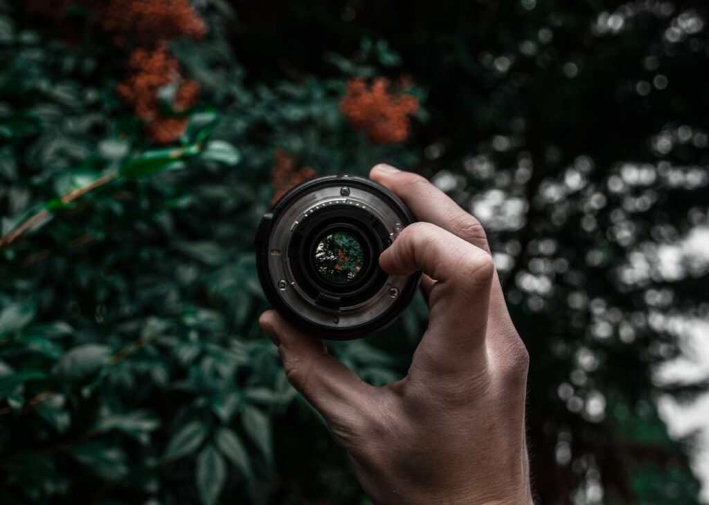 A close-up shot of a hand holding a camera lens, with leaves and blurred foliage in the background.